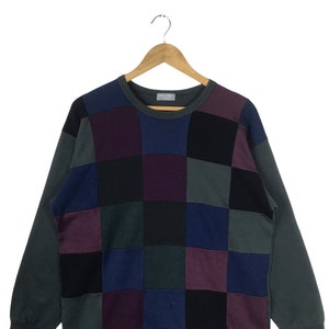 Puede incluir: Un su&eacute;ter de punto de manga larga con cuello redondo. El su&eacute;ter est&aacute; hecho de diferentes cuadrados de tela de colores, incluyendo negro, morado, azul y verde.