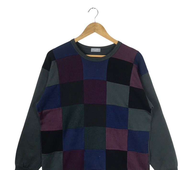 Puede incluir: Un su&eacute;ter de punto de manga larga con cuello redondo. El su&eacute;ter est&aacute; hecho de diferentes cuadrados de tela de colores, incluyendo negro, morado, azul y verde.