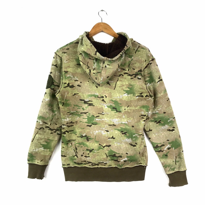 May include: A green, brown, and tan camouflage hoodie with a brown fleece lining. The hoodie has a drawstring hood and a kangaroo pocket.