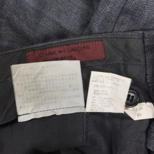 May include: A black wool trouser with a brown leather label that reads "COMME des GARCONS" and "Homme Deux". The label also includes care instructions in Japanese and English. The waist size is 80cm.