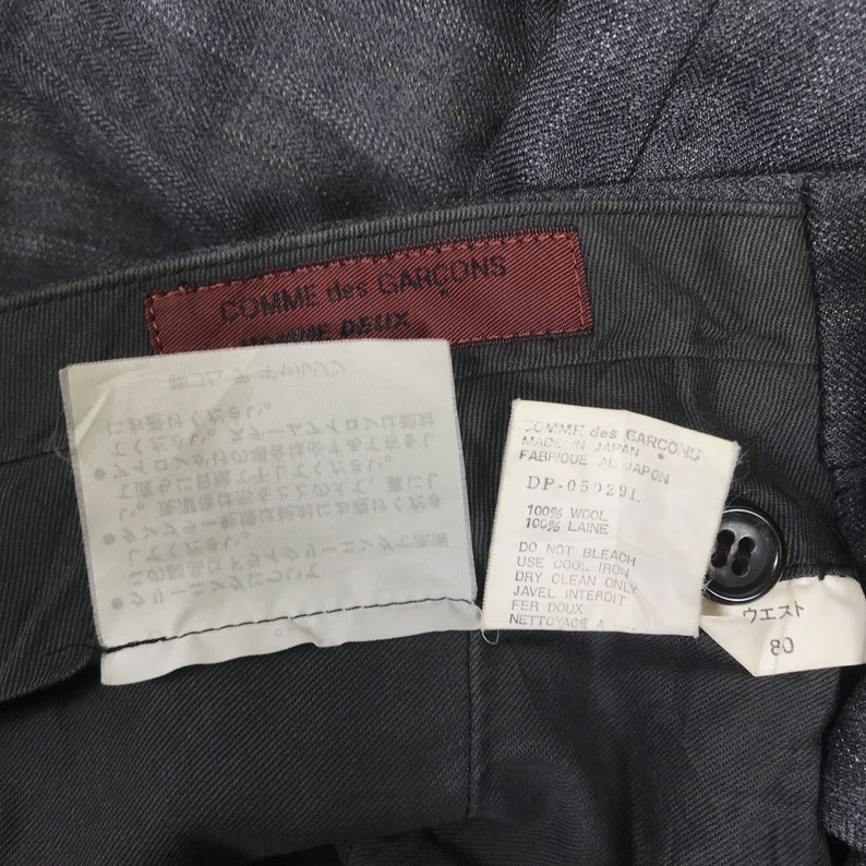 May include: A black wool trouser with a brown leather label that reads "COMME des GARCONS" and "Homme Deux". The label also includes care instructions in Japanese and English. The waist size is 80cm.