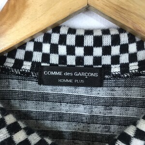 Puede incluir: Primer plano de una tela de punto a cuadros en blanco y negro con una etiqueta negra que dice "COMME des GARCONS HOMME PLUS".