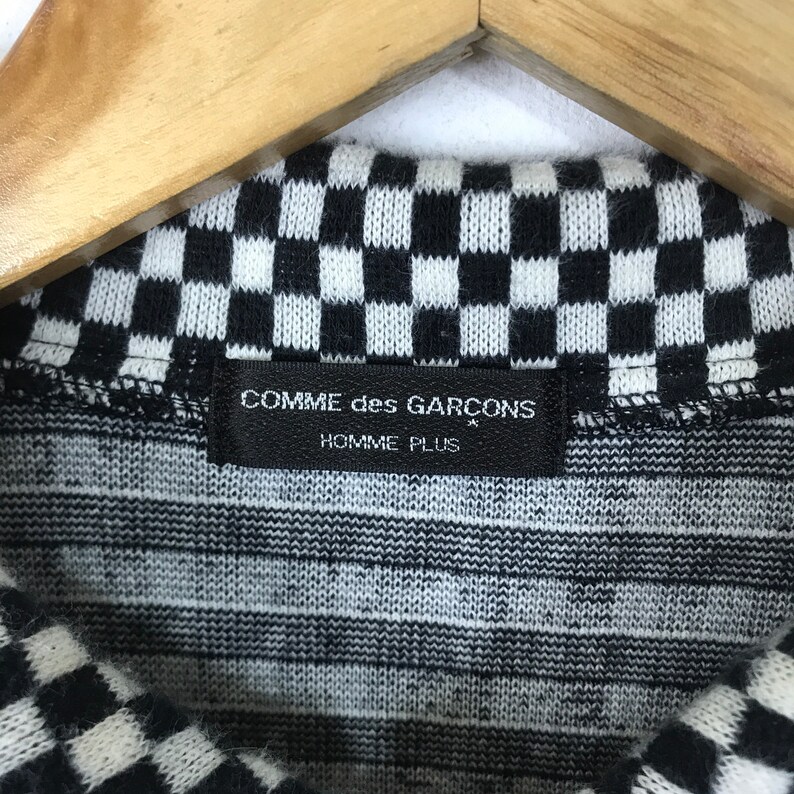 Puede incluir: Primer plano de una tela de punto a cuadros en blanco y negro con una etiqueta negra que dice "COMME des GARCONS HOMME PLUS".