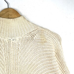 May include: A cream-colored knitted sweater with a high neck and long sleeves. The sweater is made of a soft, lightweight material and has a relaxed fit.