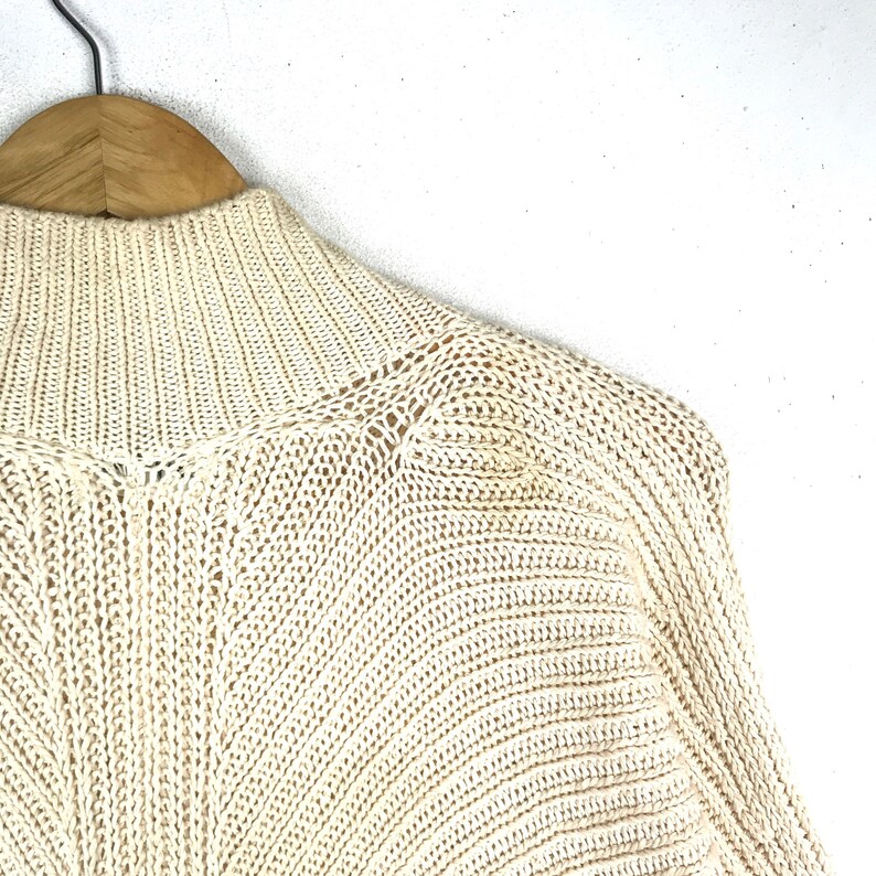 May include: A cream-colored knitted sweater with a high neck and long sleeves. The sweater is made of a soft, lightweight material and has a relaxed fit.
