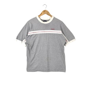 May include: A grey short-sleeved t-shirt with a white and red stripe across the chest. The shirt has the word "STUSSY" printed in red on the stripe.