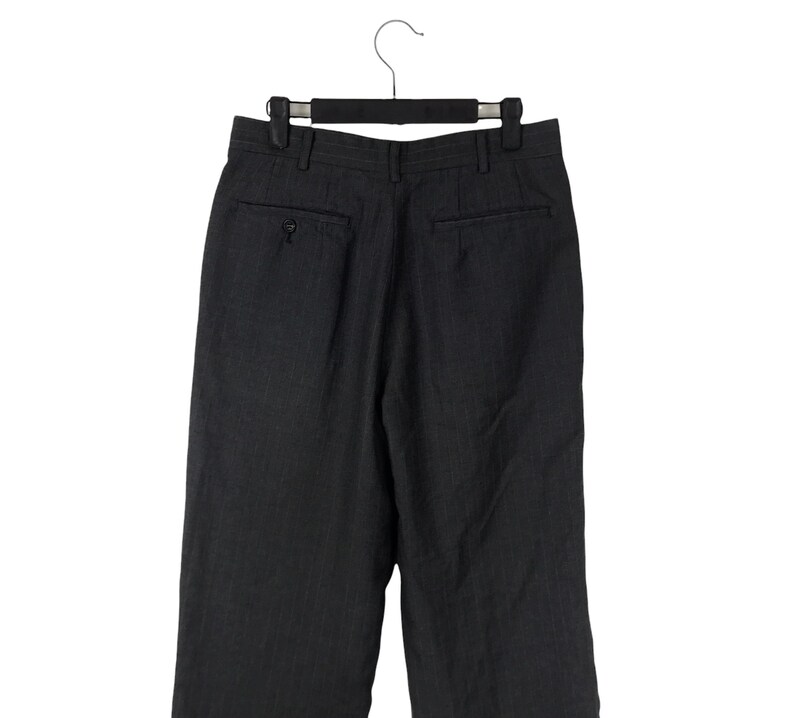 May include: Dark gray dress pants with a subtle pinstripe pattern, hanging on a black hanger. The pants have a buttoned back pocket and a classic tailored design, suitable for formal or business attire.