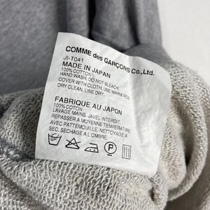 以下が含まれることがあります： グレーのスウェットシャツのクローズアップ。タグには「COMME des GAR&Ccedil;ONS Co., Ltd.」と「MADE IN JAPAN」と記載されています。洗濯表示と生地の詳細も表示されています。スウェットシャツは綿100％です。
