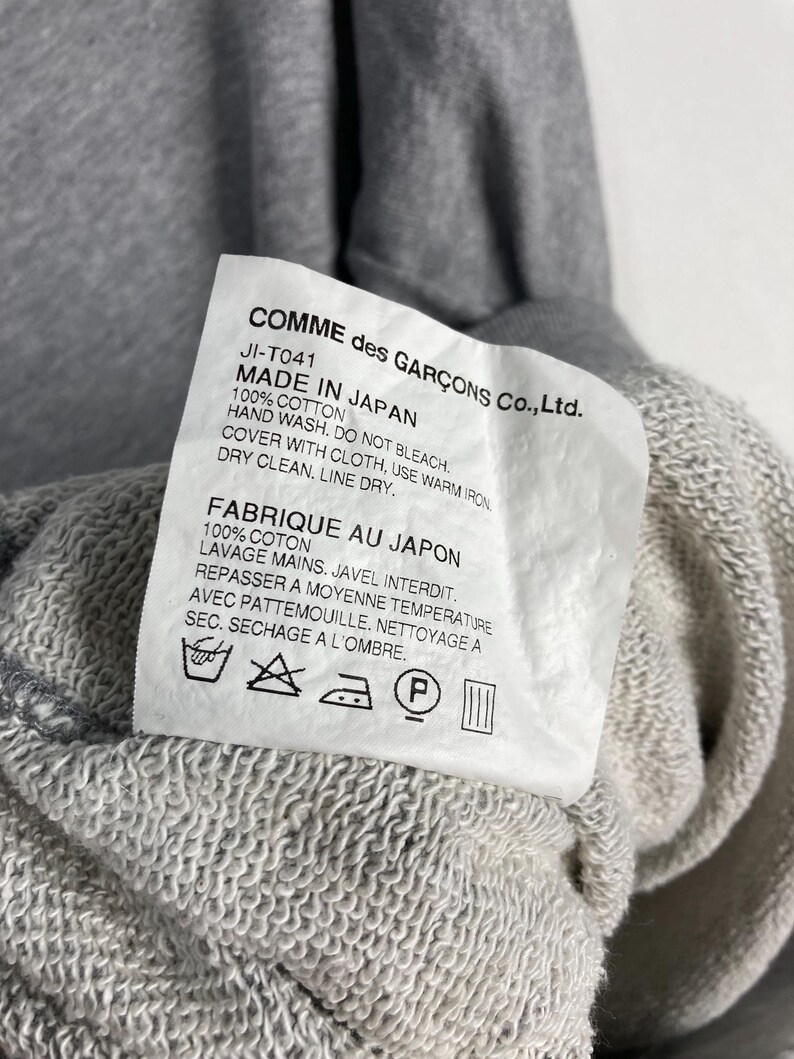 以下が含まれることがあります： グレーのスウェットシャツのクローズアップ。タグには「COMME des GAR&Ccedil;ONS Co., Ltd.」と「MADE IN JAPAN」と記載されています。洗濯表示と生地の詳細も表示されています。スウェットシャツは綿100％です。