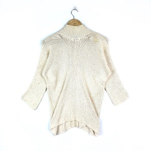 May include: A cream-colored knitted sweater with a mock neck and long sleeves. The sweater has a loose fit and a slightly asymmetrical hem.