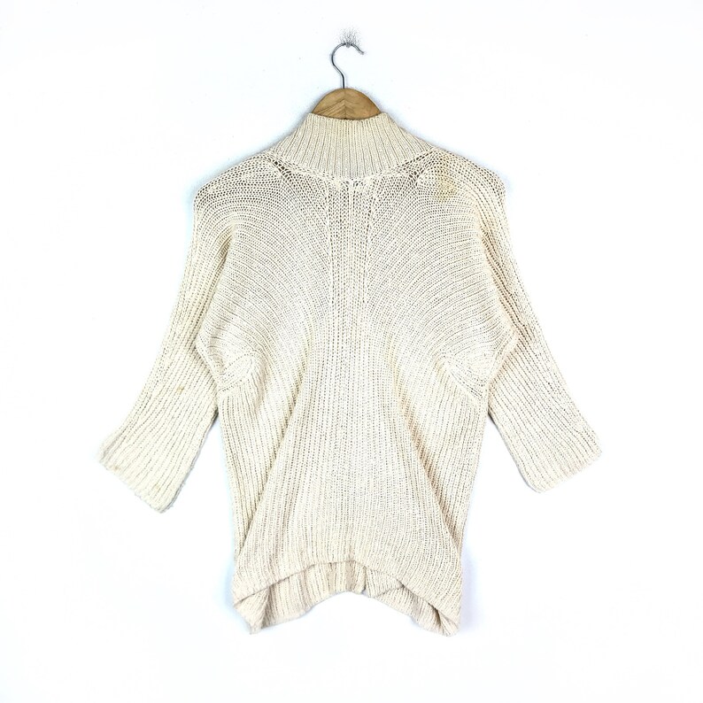 May include: A cream-colored knitted sweater with a mock neck and long sleeves. The sweater has a loose fit and a slightly asymmetrical hem.