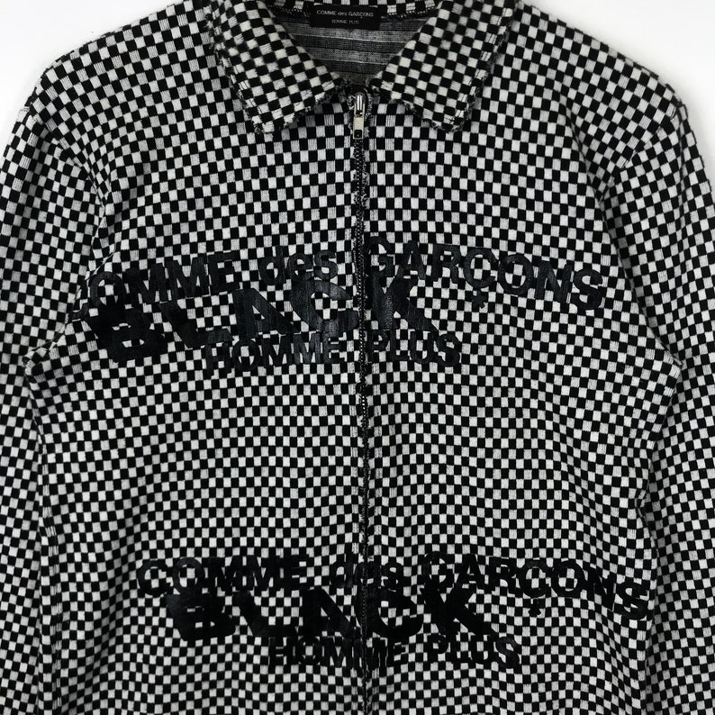 Puede incluir: Camisa de manga larga a cuadros en blanco y negro con el texto "Comme des Gar&ccedil;ons" repetido en un estampado gr&aacute;fico negro.