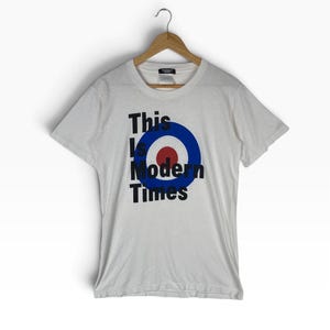 May include: A white short-sleeve t-shirt featuring a graphic of a blue and red circular target emblem. The black text "This Is Modern Times" is printed over the emblem. The shirt is displayed on a wooden hanger against a plain white background.
