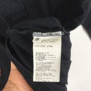 May include: Black care tag with white text detailing the care instructions for a garment. The tag reads "IQUE AU JAPON DE IN JAPAN 100% COTON COTTON NETTOYAGE A SEC (ESSENCE F) NE PAS FAIRE SECHER A LA MACHINE. A REPASSER A LA VAPEUR UNIQUEMENT FAIRE SECHER A PLAT A L'OMBRE DRY CLEAN (PETROLEUM). DO NOT TUMBLE DRY STEAM ONLY DRY FLAT IN SHADE LA SILHOUETTE PEUT PERDRE SA FORME ORIGINALE OU PEUT SE RETRECIR LORS DU LAVAGE DOVESTIQUE THE ORIGINAL SHAPE COULD SLIGHTLY COME DOWN OR SHRINK BY HOME WASHING NE PA FAIRE SECHER A MACHINE DO NOT TUMBLE DRY"