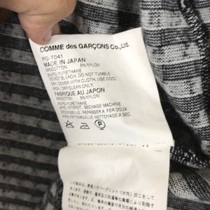 Puede incluir: Una etiqueta blanca con texto negro sobre una tela rayada en blanco y negro. La etiqueta dice "COMME des GAR&Ccedil;ONS Co., Ltd." e incluye instrucciones de cuidado en ingl&eacute;s y franc&eacute;s. La etiqueta tambi&eacute;n incluye un n&uacute;mero de tama&ntilde;o y estilo.