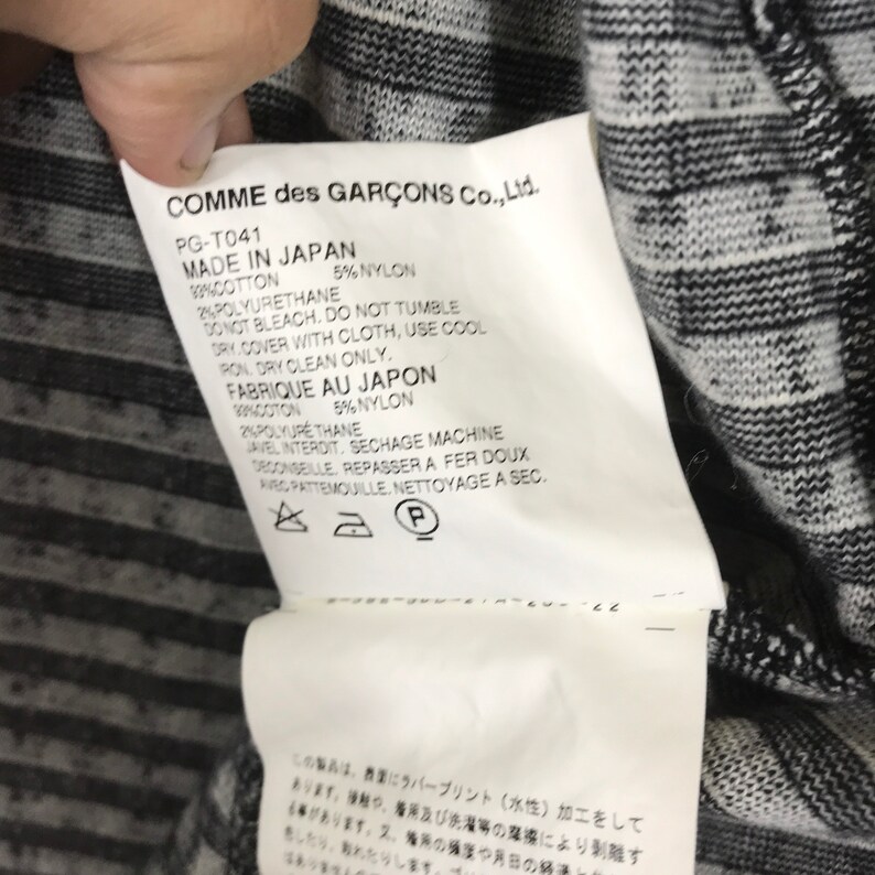 Puede incluir: Una etiqueta blanca con texto negro sobre una tela rayada en blanco y negro. La etiqueta dice "COMME des GAR&Ccedil;ONS Co., Ltd." e incluye instrucciones de cuidado en ingl&eacute;s y franc&eacute;s. La etiqueta tambi&eacute;n incluye un n&uacute;mero de tama&ntilde;o y estilo.