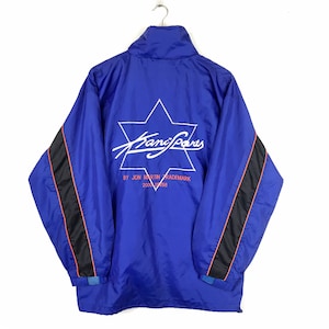 May include: A blue windbreaker jacket with black and red stripes on the sleeves. The back of the jacket features a white star with the text "Kani Sports" in white and red lettering. The text "By Jon Martin Trademark 2000 32158" is printed below the star.
