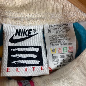K&ouml;nnte beinhalten: Nahaufnahme eines cremefarbenen Nike Elite Jackenetiketts. Das Etikett zeigt das Nike-Logo, Produktinformationen und Waschanweisungen. Der Rei&szlig;verschluss der Jacke ist unten sichtbar, mit einem rosa und grauen Design.