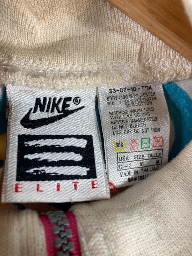 K&ouml;nnte beinhalten: Nahaufnahme eines cremefarbenen Nike Elite Jackenetiketts. Das Etikett zeigt das Nike-Logo, Produktinformationen und Waschanweisungen. Der Rei&szlig;verschluss der Jacke ist unten sichtbar, mit einem rosa und grauen Design.