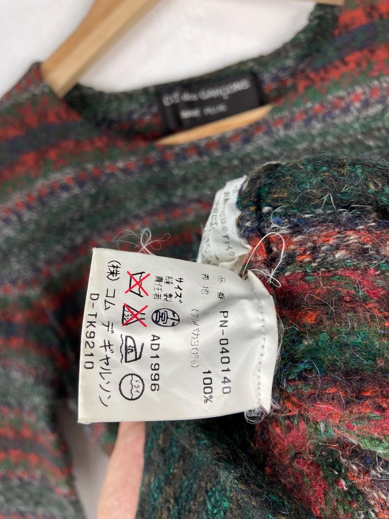 May include: A white tag with black text detailing the care instructions for a garment. The tag reads "(株) コムデギャルソン D-TK9210", "ドライ", "AD1996", "品番 PN-040140", "表地 100% (アルパカ30%)", and "おかには必ずェイク)".