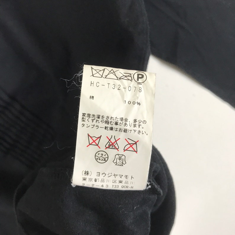 May include: A black clothing care label with white text and symbols. The label includes the text "HC-T32-078", "綿 100%", "(株) ヨウジヤマモト", "東京都品川区東品川 2-2-43 T33 OCR-H", and several care symbols.