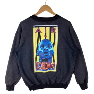 May include: A grey sweatshirt with a black crew neck and a graphic print on the back. The graphic features a yellow rectangle with the text "AIR BY JORDAN" in red, blue, and white. A blue and white image of Michael Jordan is in the centre of the rectangle.