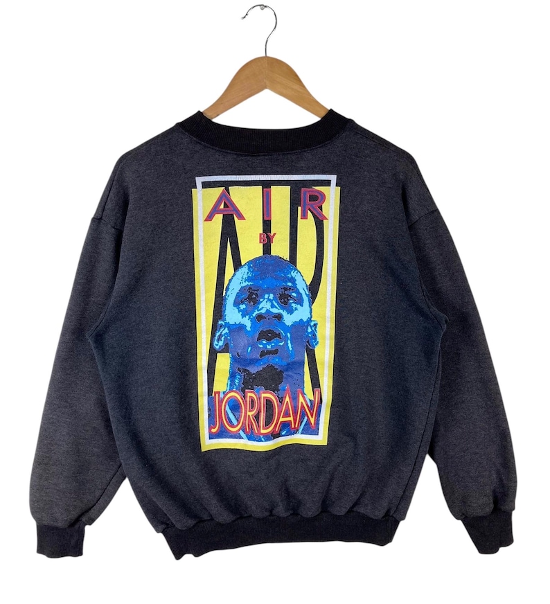 May include: A grey sweatshirt with a black crew neck and a graphic print on the back. The graphic features a yellow rectangle with the text "AIR BY JORDAN" in red, blue, and white. A blue and white image of Michael Jordan is in the centre of the rectangle.