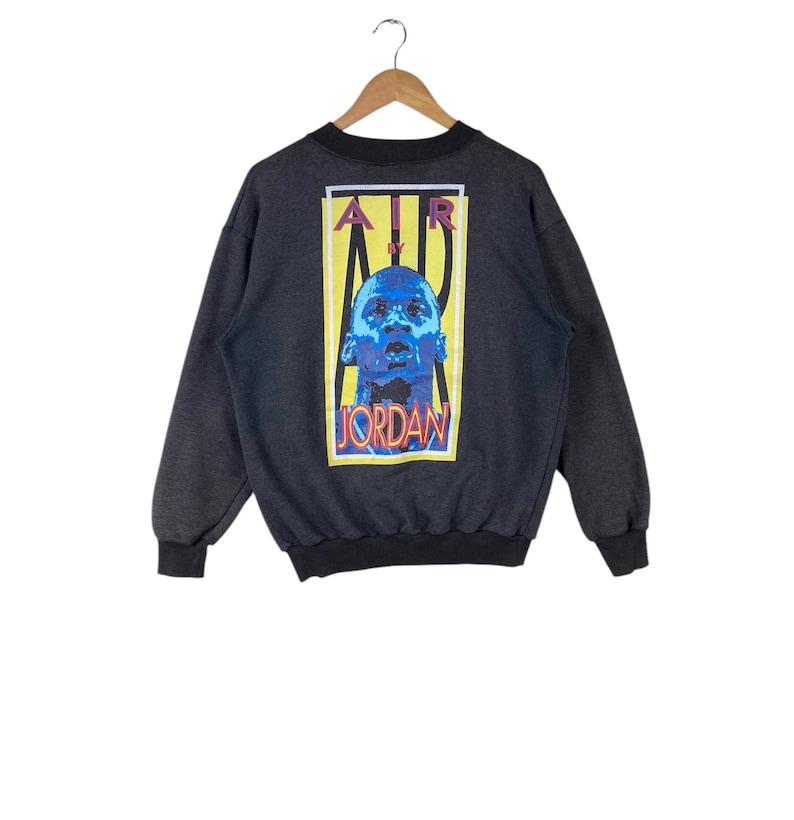 May include: A dark grey sweatshirt with a large graphic print on the back. The graphic features a yellow rectangle with the text "AIR BY JORDAN" in red, black, and white. Inside the rectangle is a blue and white image of Michael Jordan.