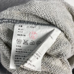 以下が含まれることがあります： グレーのスウェットシャツのクローズアップ。タグには、製品コード「JI-T041」や洗濯表示など、日本語のテキストが表示されています。生地は、テクスチャード加工されたコットン混紡のようです。