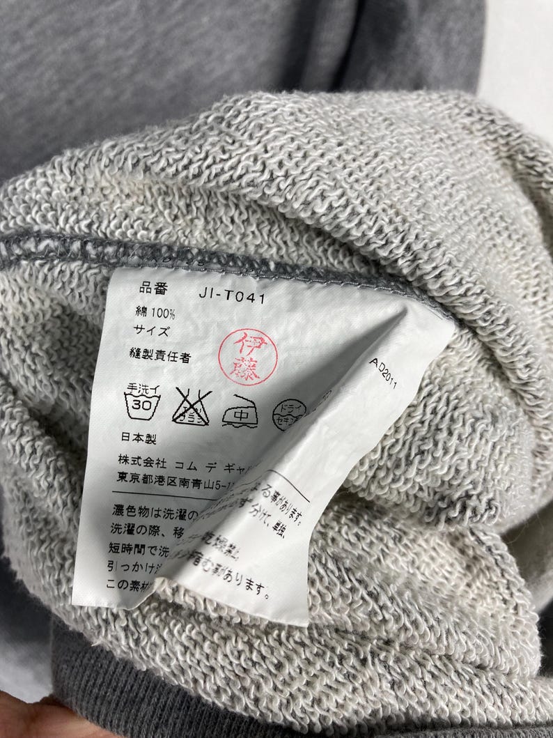 以下が含まれることがあります： グレーのスウェットシャツのクローズアップ。タグには、製品コード「JI-T041」や洗濯表示など、日本語のテキストが表示されています。生地は、テクスチャード加工されたコットン混紡のようです。