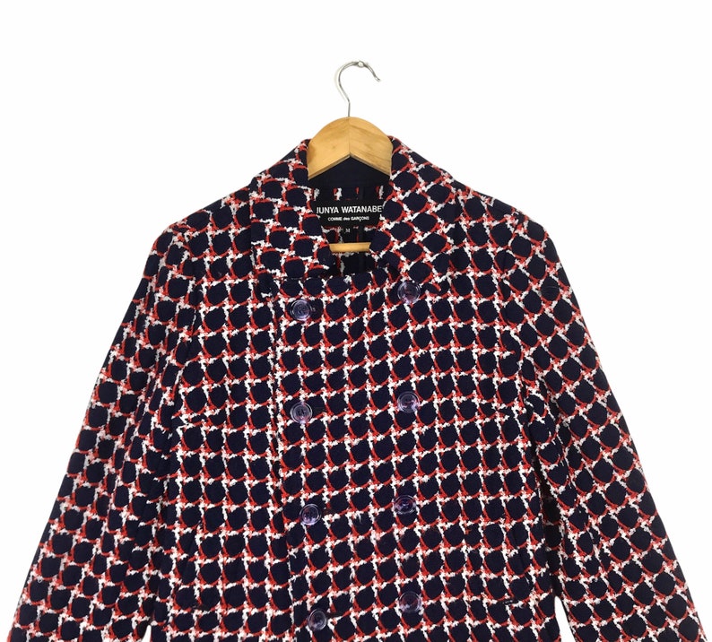 May include: A double-breasted navy blue jacket with a geometric pattern in white and red. The jacket is hanging on a wooden hanger. The label reads "JUNYA WATANABE COMME des GAR&Ccedil;ONS".