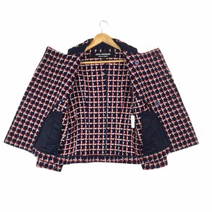 May include: A navy blue and white checkered jacket with red accents, hanging on a wooden hanger. The jacket has two front pockets and a label that reads "JUNYA WATANABE COMME des GAR&Ccedil;ONS".