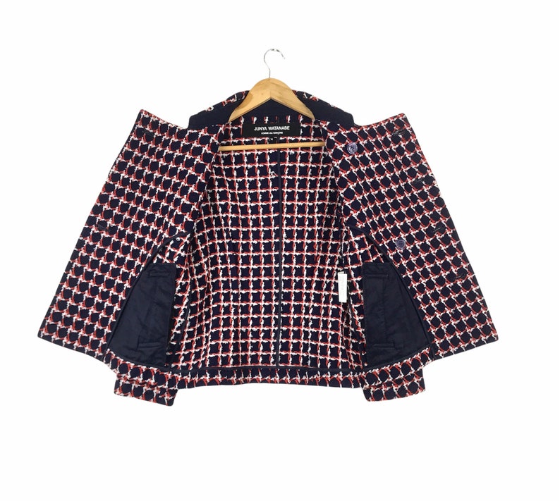 May include: A navy blue and white checkered jacket with red accents, hanging on a wooden hanger. The jacket has two front pockets and a label that reads "JUNYA WATANABE COMME des GAR&Ccedil;ONS".