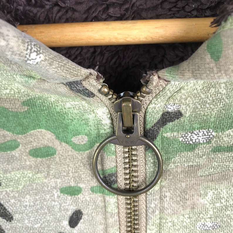 May include: Close-up of a zipper on a camouflage hoodie. The zipper has a large, round pull with the word "YKK" inscribed on it.