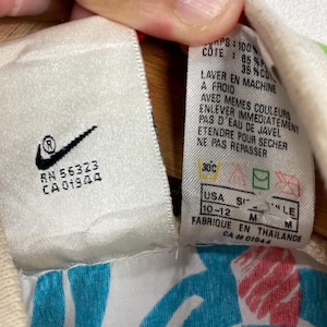 K&ouml;nnte beinhalten: Nahaufnahme eines wei&szlig;en Etiketts mit dem Nike-Logo und Text. Das Etikett enth&auml;lt Waschanweisungen auf Franz&ouml;sisch und Gr&ouml;&szlig;enangaben, einschlie&szlig;lich der US-Gr&ouml;&szlig;e 10-12. Das Etikett ist an einem Kleidungsst&uuml;ck mit blauem und wei&szlig;em Muster befestigt.