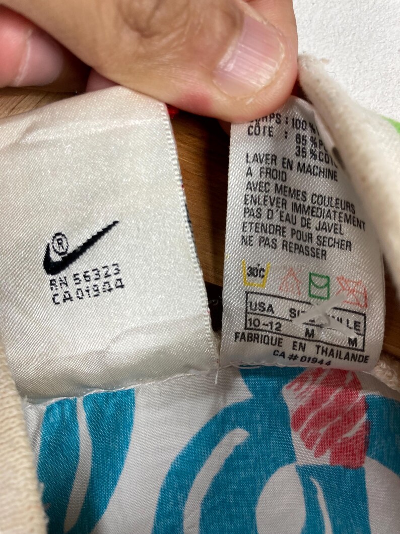 K&ouml;nnte beinhalten: Nahaufnahme eines wei&szlig;en Etiketts mit dem Nike-Logo und Text. Das Etikett enth&auml;lt Waschanweisungen auf Franz&ouml;sisch und Gr&ouml;&szlig;enangaben, einschlie&szlig;lich der US-Gr&ouml;&szlig;e 10-12. Das Etikett ist an einem Kleidungsst&uuml;ck mit blauem und wei&szlig;em Muster befestigt.