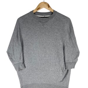 以下が含まれることがあります： 木製のハンガーに吊るされた、3/4スリーブのヘザーグレーのスウェットシャツ。クルーネックのスウェットシャツは、リブ編みの襟、袖口、裾が付いています。袖は3/4丈です。この衣服は、柔らかく快適な素材で作られています。
