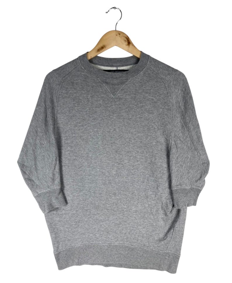 以下が含まれることがあります： 木製のハンガーに吊るされた、3/4スリーブのヘザーグレーのスウェットシャツ。クルーネックのスウェットシャツは、リブ編みの襟、袖口、裾が付いています。袖は3/4丈です。この衣服は、柔らかく快適な素材で作られています。