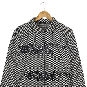 Puede incluir: Chaqueta con cremallera de cuadros blancos y negros con el texto "Comme des Gar&ccedil;ons Black" impreso en la parte delantera.