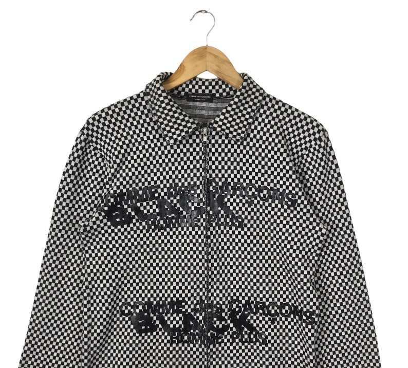 Puede incluir: Chaqueta con cremallera de cuadros blancos y negros con el texto "Comme des Gar&ccedil;ons Black" impreso en la parte delantera.