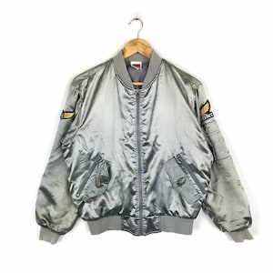 May include: A silver satin bomber jacket with a zip closure and two front pockets. The jacket has a yellow and black embroidered patch on the left sleeve and a yellow and black embroidered patch on the right sleeve that says "HONDA".