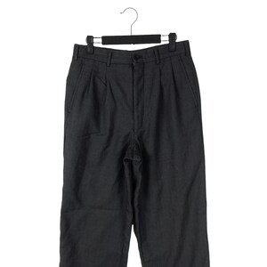 May include: Dark gray pleated trousers hanging on a black hanger. The pants have a button closure and pockets. The fabric appears to have a subtle vertical stripe pattern. The trousers are displayed against a white background.