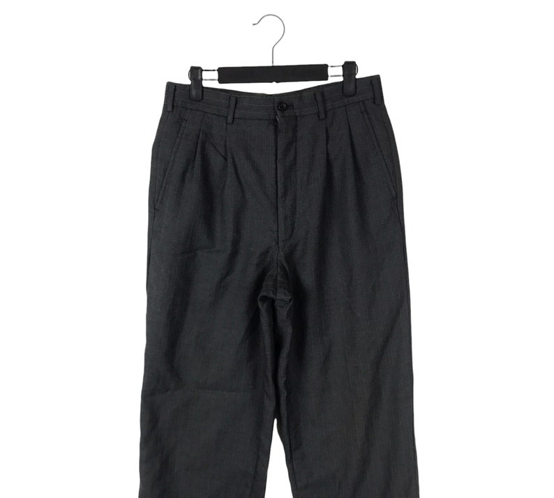 May include: Dark gray pleated trousers hanging on a black hanger. The pants have a button closure and pockets. The fabric appears to have a subtle vertical stripe pattern. The trousers are displayed against a white background.