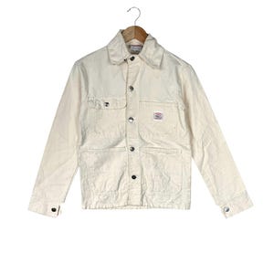 May include: Cream-colored work jacket with a button-up front, two chest pockets, and two lower pockets. The jacket has a collar and buttoned cuffs. A small label is visible on the right chest pocket.