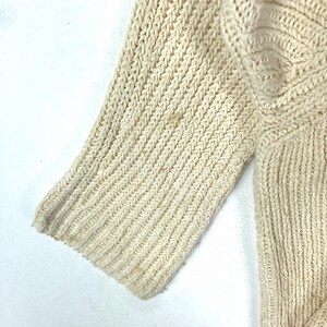 May include: Close-up of a cream-colored knitted sweater sleeve. The fabric is a ribbed knit with a fine texture.