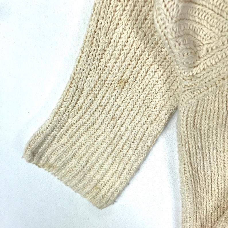 May include: Close-up of a cream-colored knitted sweater sleeve. The fabric is a ribbed knit with a fine texture.