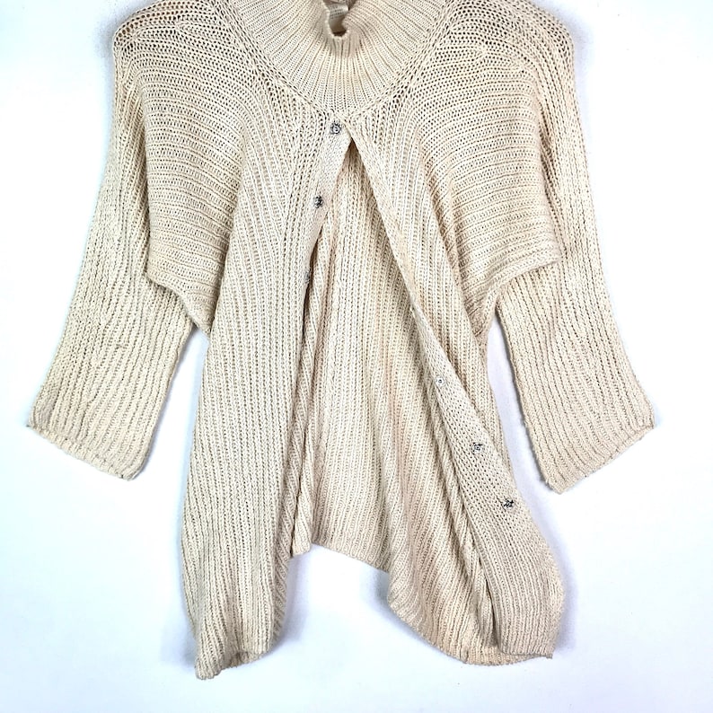 May include: A cream-colored knitted cardigan with a button closure and a slightly asymmetrical hem.