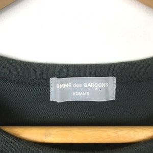 Puede incluir: Una camiseta gris oscuro con una etiqueta blanca que dice "Comme des Gar&ccedil;ons Homme"