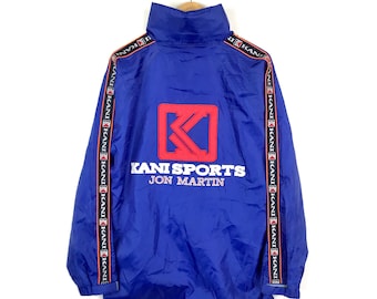 Pode incluir: Casaco corta-vento de nylon azul com logotipo KANI SPORTS JON MARTIN vermelho e branco e letras KANI vermelhas e brancas nas mangas.