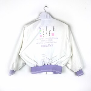 May include: White windbreaker jacket with a purple zipper and waistband. The back of the jacket has a colorful logo that says "ELLE SSE" and the text "FOR CHI VIVE IL TENNIS IN TERMINE DI SPORT ATTIVO IN MODO GIOVANE DIVERTENTE PERUGIA ITALY".